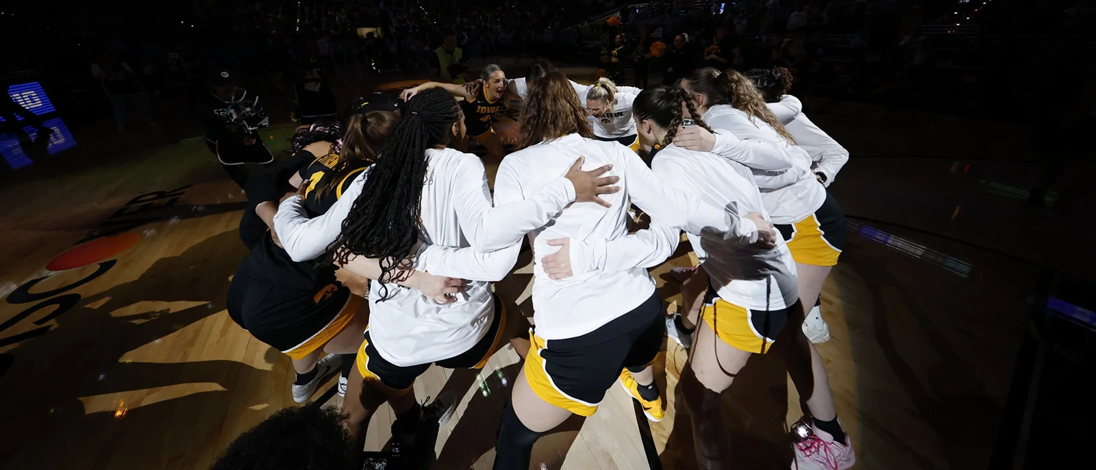 Iowa women's basketball players in white warmups embracing in a pregame huddle