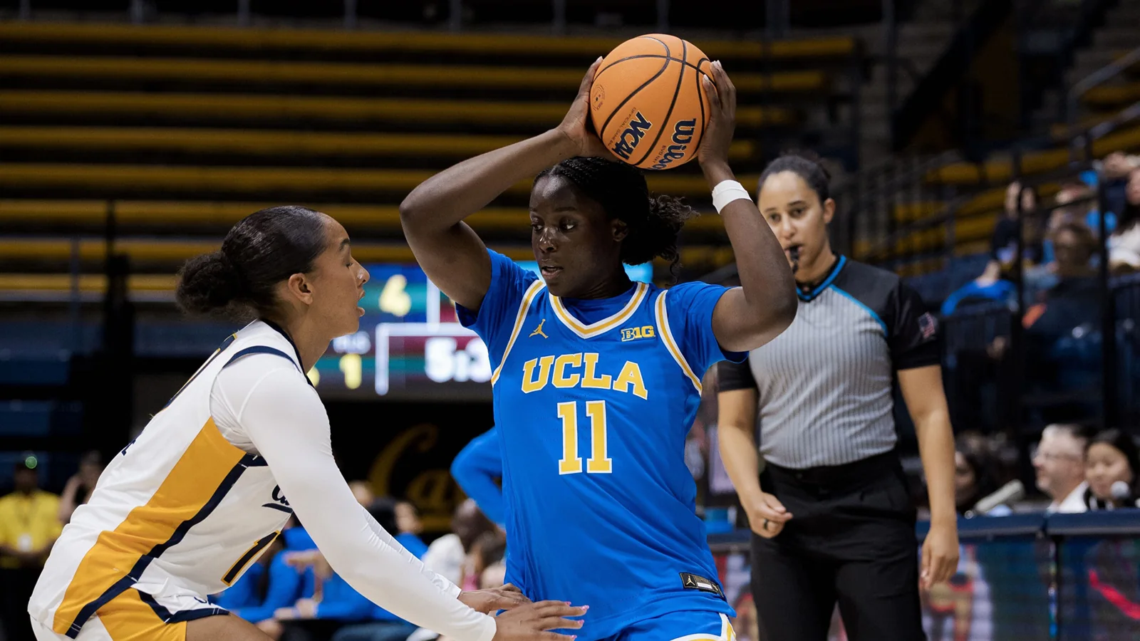 UCLA women's basketball player in a blue number 11 jersey holding the ball overhead as a Cal defender guards her
