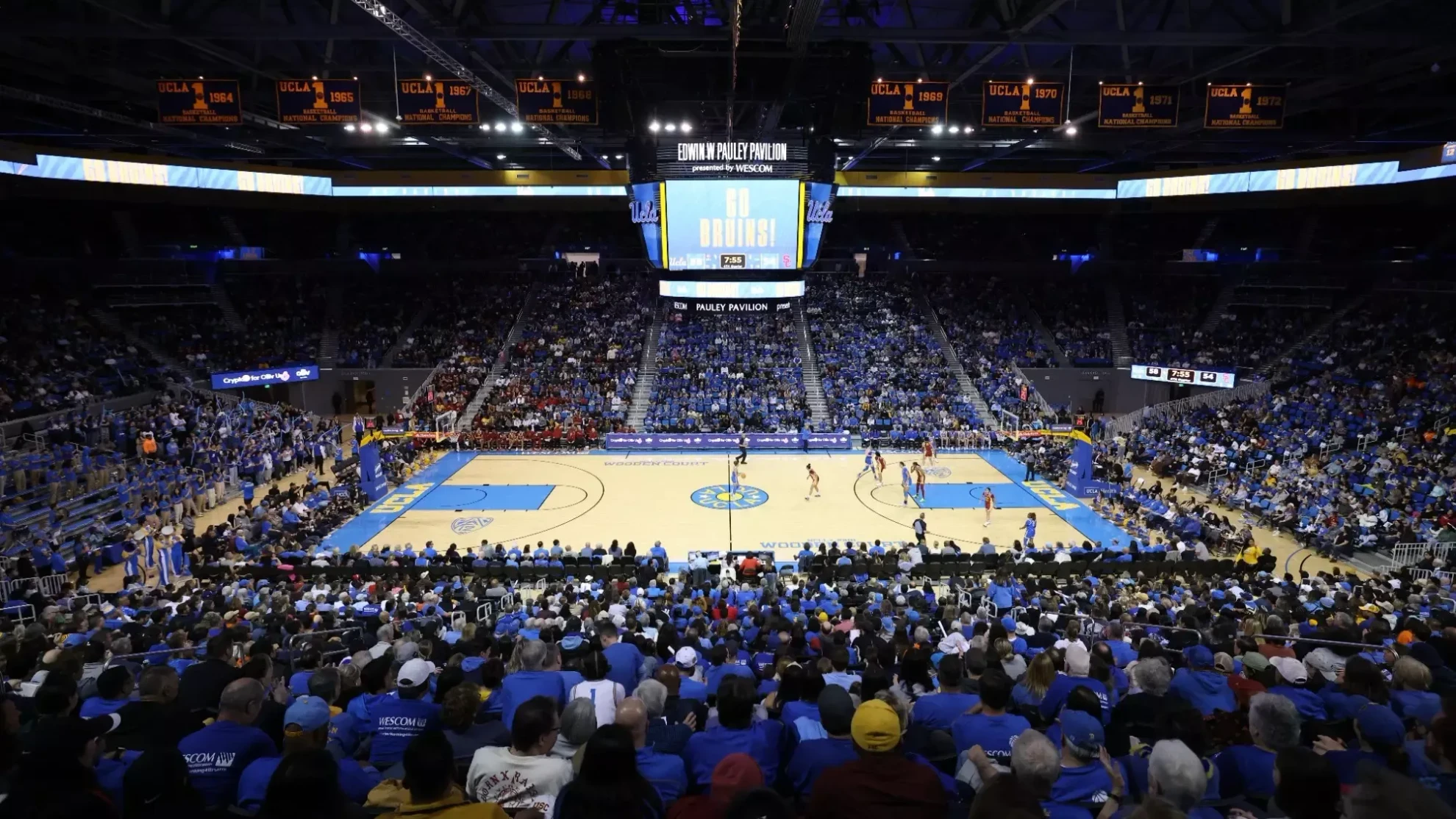 Wide interior view of a packed Pauley Pavilion during a UCLA women's basketball game, with UCLA men's basketball national championship banners from the 1960s and 1970s hanging above the court